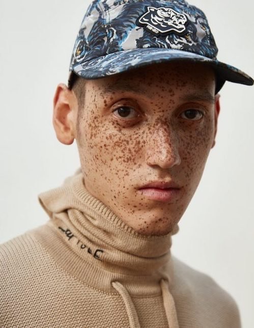 Portrait of a young adult male with freckles wearing a patterned baseball cap and a tan turtleneck sweater, in a studio setting.