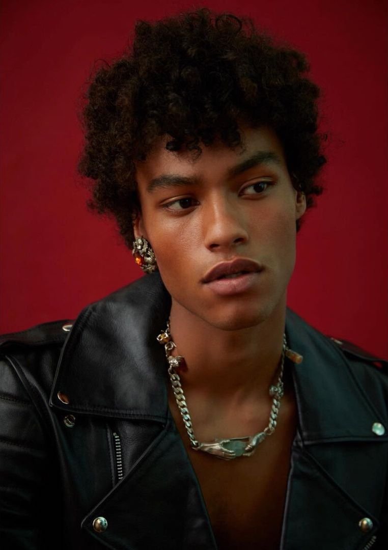 Close-up portrait of a young adult male with afro, wearing a black leather jacket, silver necklace, and earring against a red backdrop.