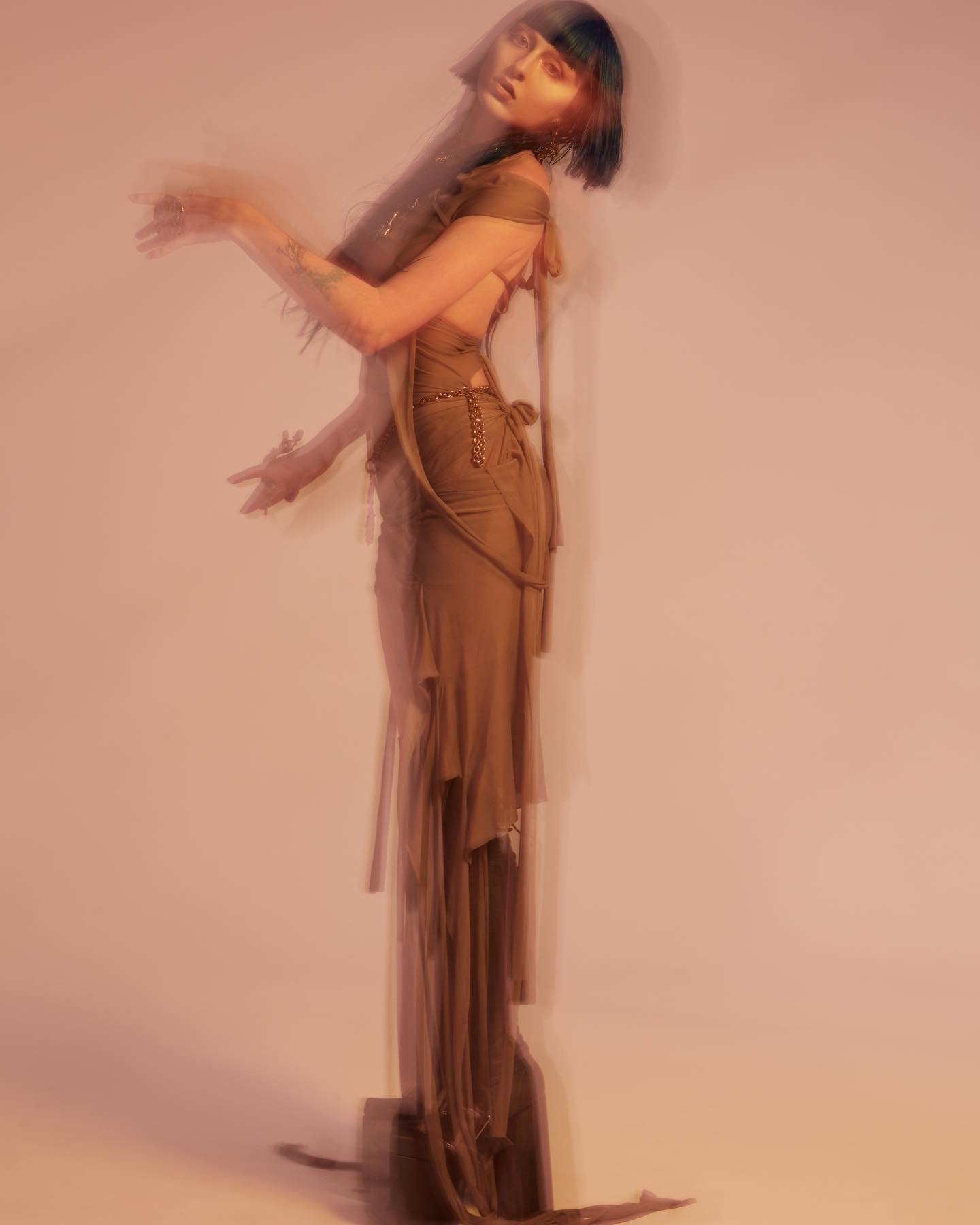 Woman with dark hair poses in a flowing dress in a blurred artistic style against a light pink background, capturing a dreamy aesthetic.