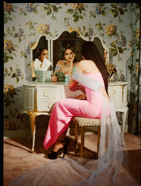 Woman in pink jumpsuit seated at vanity with floral wallpaper, looking in mirror, with tulle fabric and black platform heels.