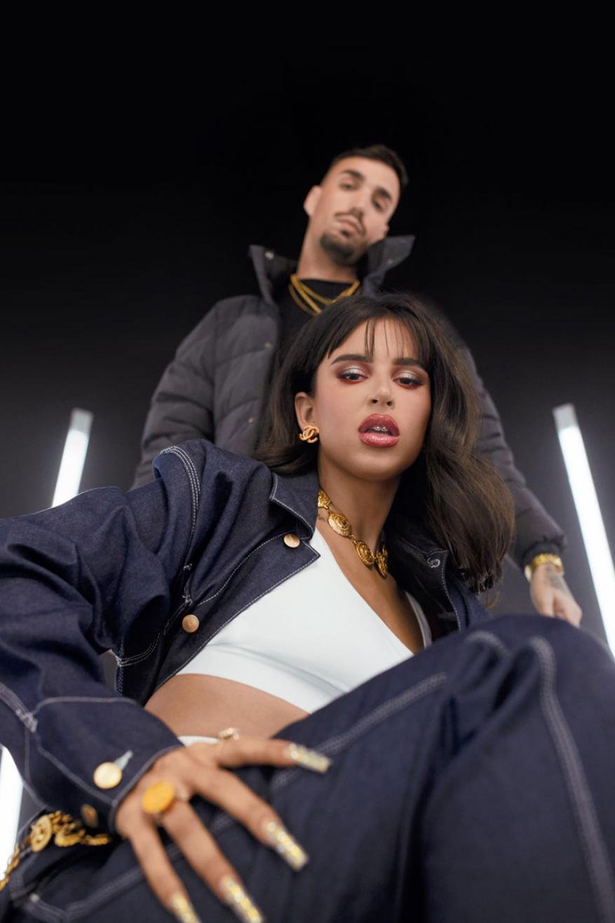 A woman in a dark denim jacket and white top with a man in a black puffer jacket in a studio with vertical light bars.
