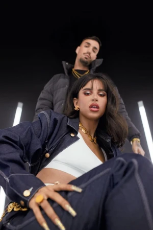 A woman in a dark denim jacket and white top with a man in a black puffer jacket in a studio with vertical light bars.