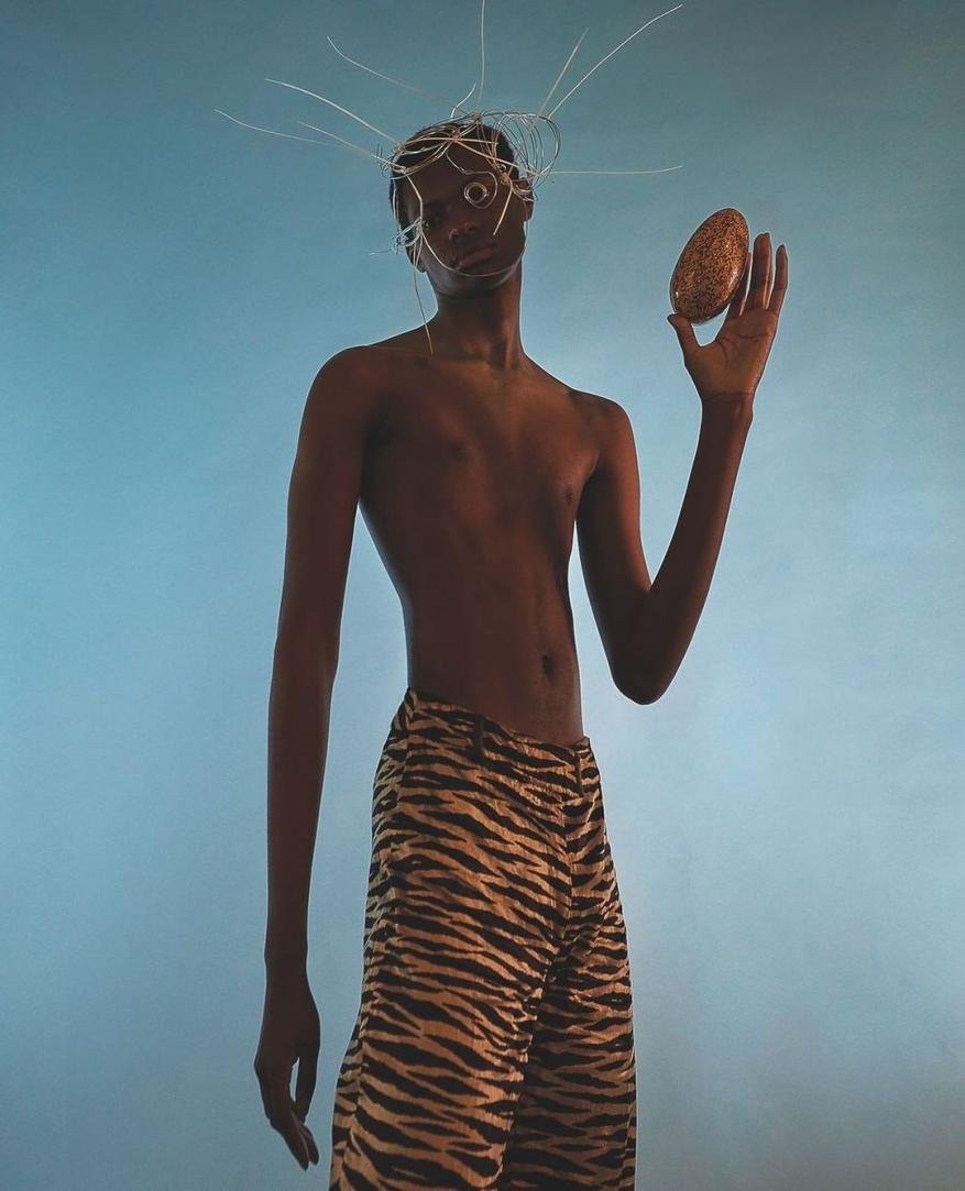 Dark-skinned person stands in front of blue backdrop, wearing tiger-striped pants and holding oval object, silver wire headpiece.
