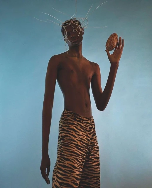 Dark-skinned person stands in front of blue backdrop, wearing tiger-striped pants and holding oval object, silver wire headpiece.