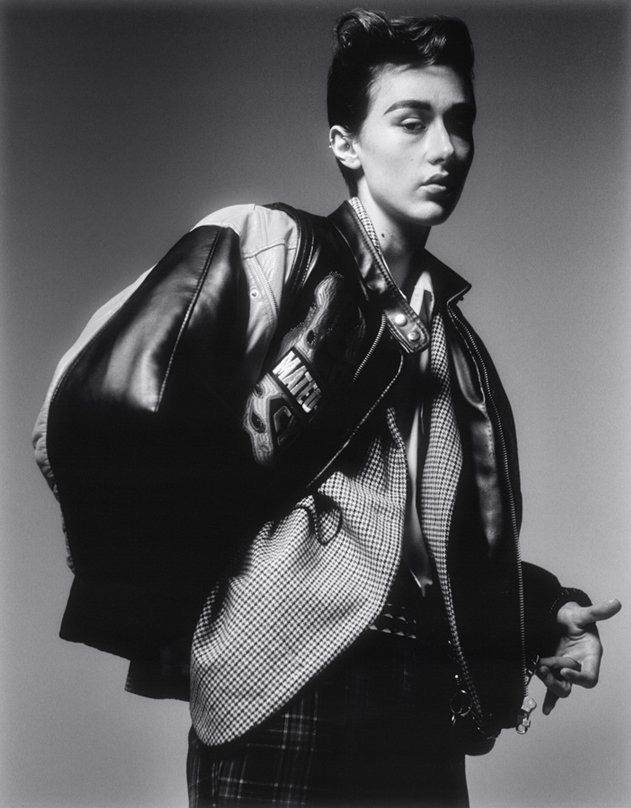 Monochrome portrait of a person in a leather bomber jacket draped over shoulders and patterned blazer, shot against a neutral studio background.