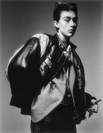 Monochrome portrait of a person in a leather bomber jacket draped over shoulders and patterned blazer, shot against a neutral studio background.