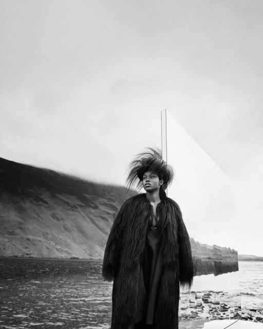 Fashion shot of a woman in a fur coat standing in a rocky, outdoor environment with water, hills, and a cloudy sky in monochrome.