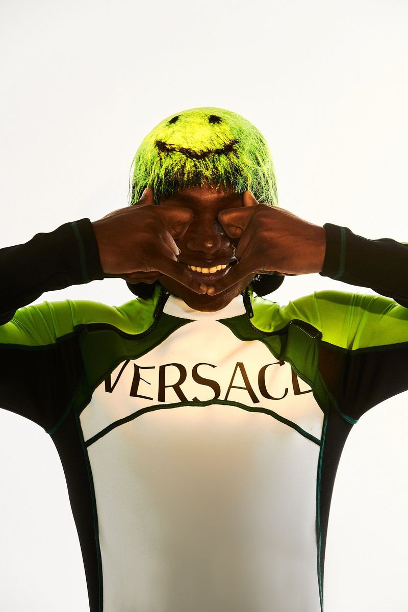 Person with green hair and a smiley face drawn on their head, wearing a Versace top, making shapes over their eyes with their hands.