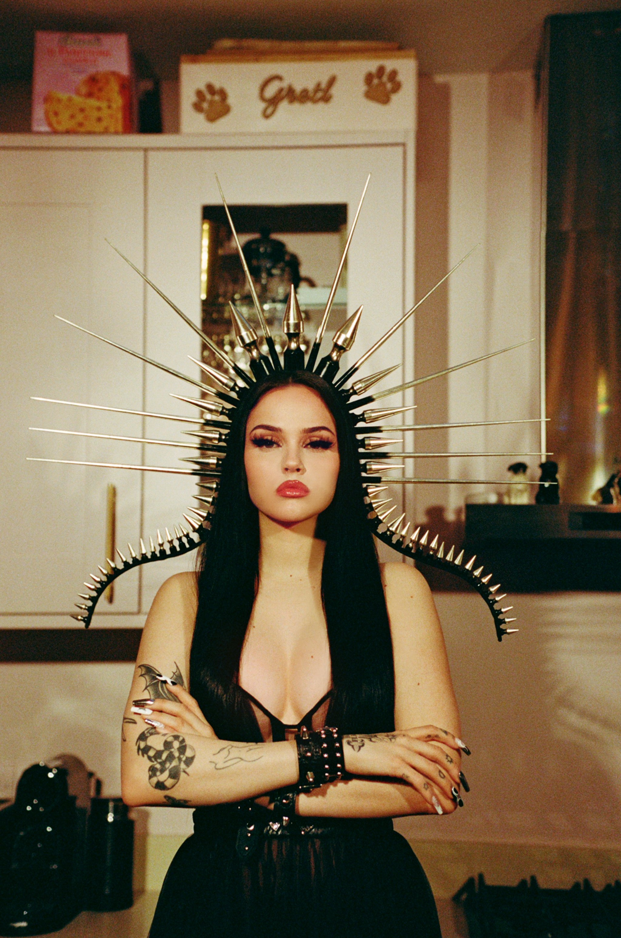 A young adult woman stands indoors wearing an elaborate gold headdress, black clothing, and arm tattoos, posing in a kitchen setting.