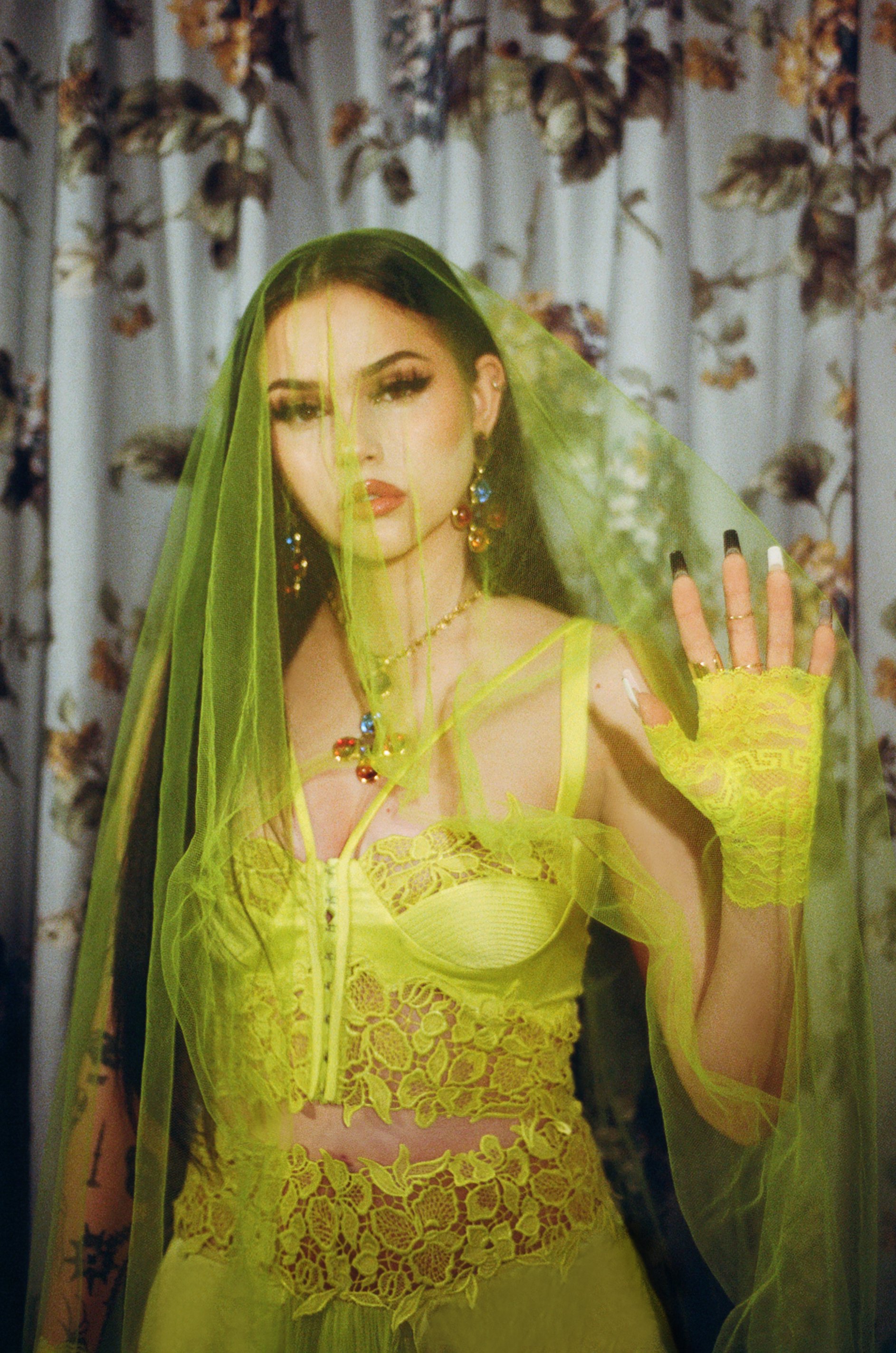 A woman poses in a vibrant yellow lace top, sheer skirt, and a flowing veil against a floral backdrop. Her long black nails are visible.
