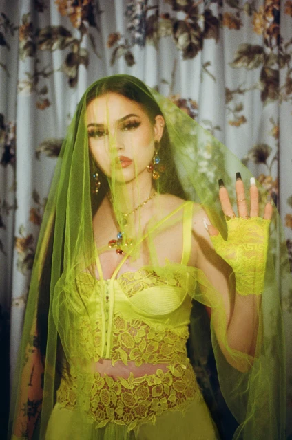 A woman poses in a vibrant yellow lace top, sheer skirt, and a flowing veil against a floral backdrop. Her long black nails are visible.