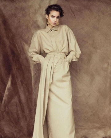 Model standing against a textured brown backdrop wearing a long beige belted shirt dress and statement earrings.