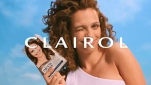 Woman winks while holding Clairol hair color box against a blue sky. She has curly hair and wears a white top.