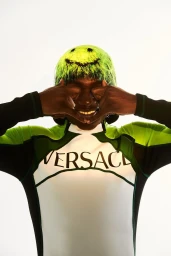 Model wearing a bright green smiley face hat and Versace top with hands framing face against a white background.