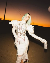 Woman with mint-green bob hair wearing a white satin dress and harness standing outdoors at sunset.