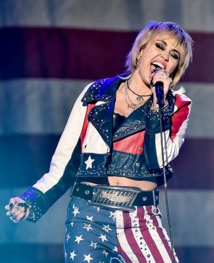 Miley Cyrus sings into a microphone on stage, wearing a jacket and pants with an American flag design in a concert setting.