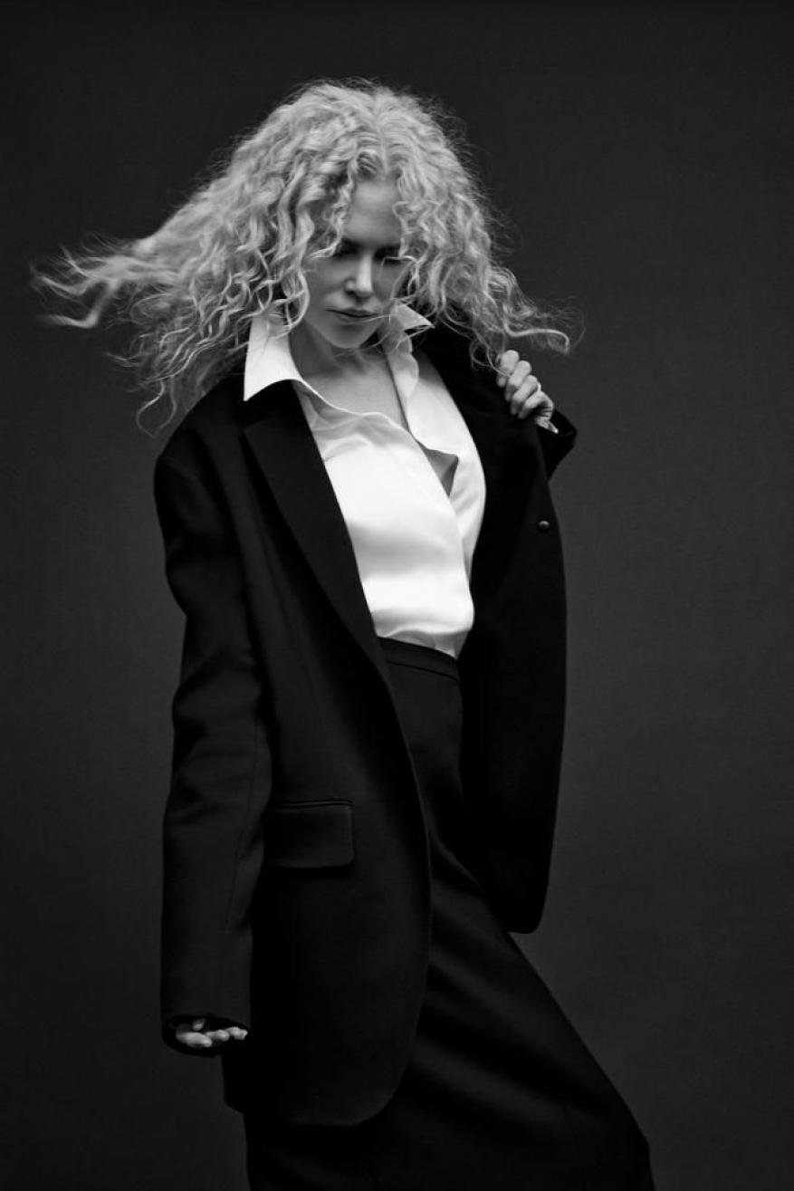 Black and white portrait of a stylish woman with curly blonde hair, wearing a white blouse and oversized dark blazer in a studio.