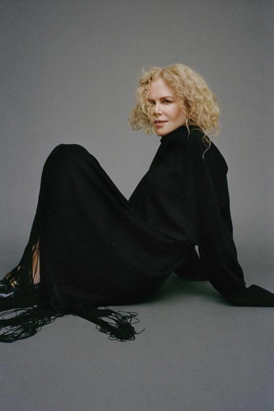 Indoor studio shot of a blonde woman in a black turtleneck dress, seated on a gray backdrop, looking at the camera.
