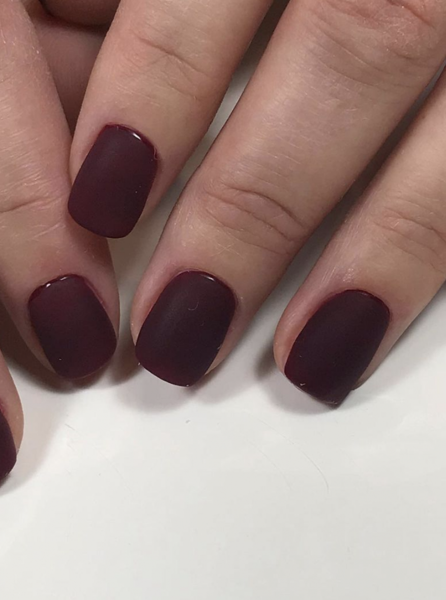 Close-up of a woman's manicured fingers featuring matte dark red nails against a white background, showcasing beauty and nail care.