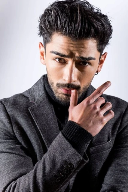 Stylish male model in gray coat and black turtleneck poses in a studio portrait with a finger to his lips, conveying a sense of secrecy.