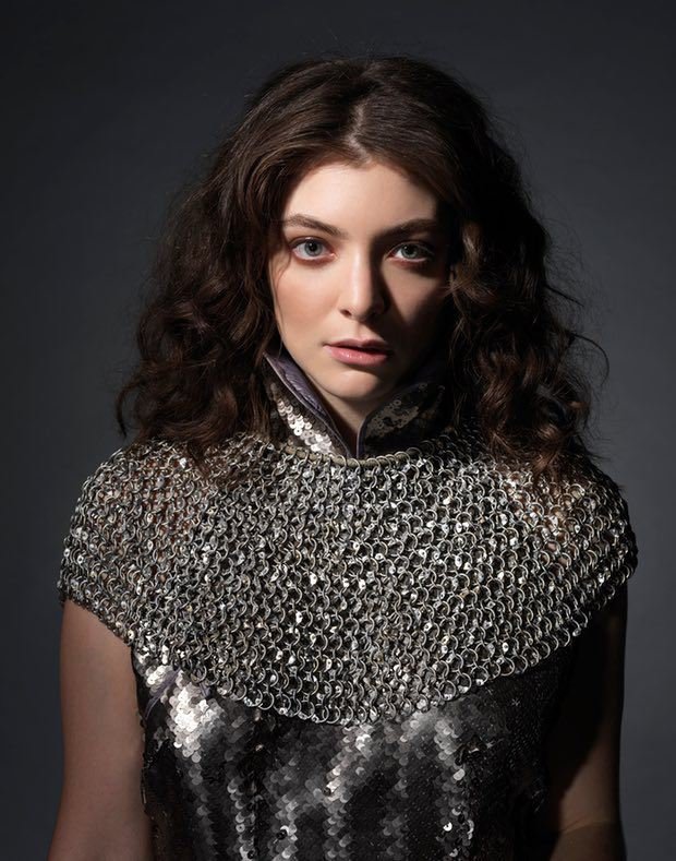 Close-up portrait of a female with shoulder-length curly brown hair wearing a metallic chainmail top against a dark background.