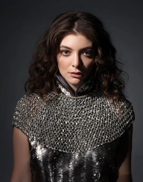 Close-up portrait of a female with shoulder-length curly brown hair wearing a metallic chainmail top against a dark background.