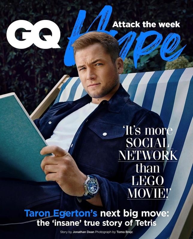 GQ magazine cover featuring Taron Egerton reclining on a striped lounge chair, reading a book, wearing a navy jacket with a wristwatch.