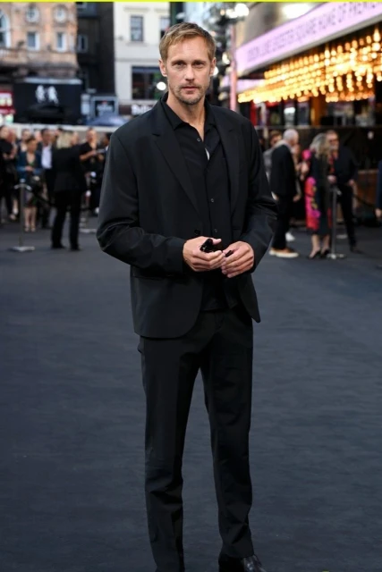 Alexander Skarsgård in a dark suit on a red carpet at an event. The man is facing forward with a neutral expression, full shot.
