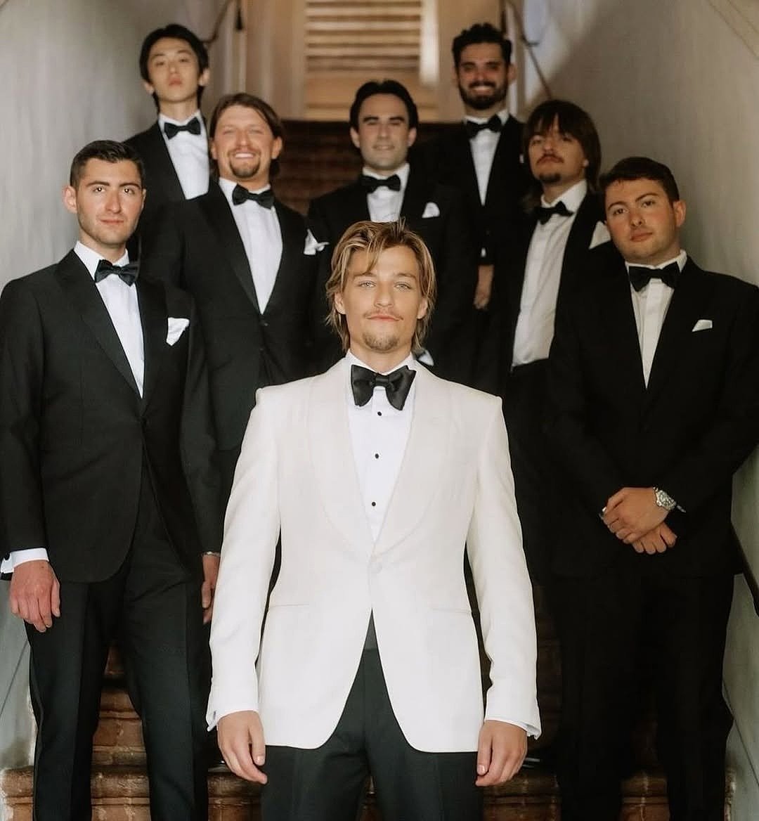 A group of eight men in tuxedos standing on stairs, posing for a portrait, indoors. One man in a cream colored tuxedo stands in front.