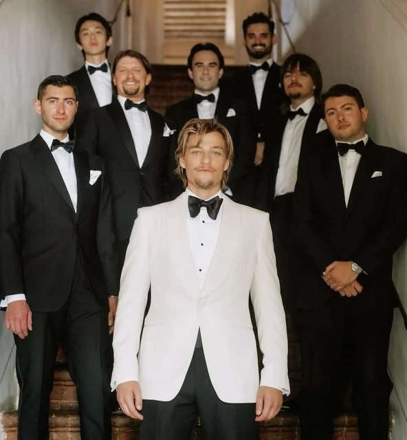 A group of eight men in tuxedos standing on stairs, posing for a portrait, indoors. One man in a cream colored tuxedo stands in front.