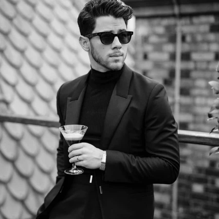 Monochrome portrait of Nick Jonas wearing sunglasses and a black blazer, holding a cocktail glass on a rooftop terrace.