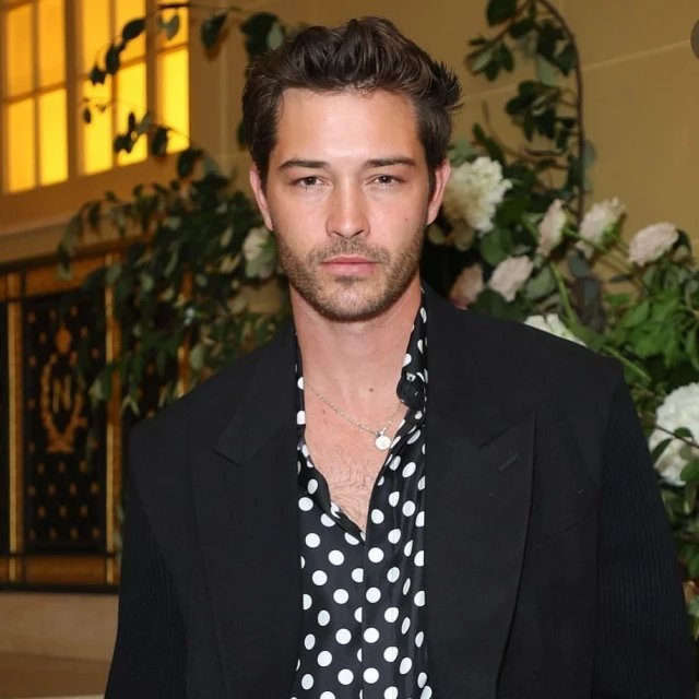 Adult male in a polka-dot shirt and black suit jacket poses indoors with floral arrangements. Fashion and celebrity event photography.