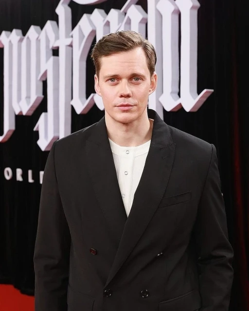 Close-up of fair-skinned adult male in black double-breasted suit, wearing white henley, in front of red backdrop with white letters.
