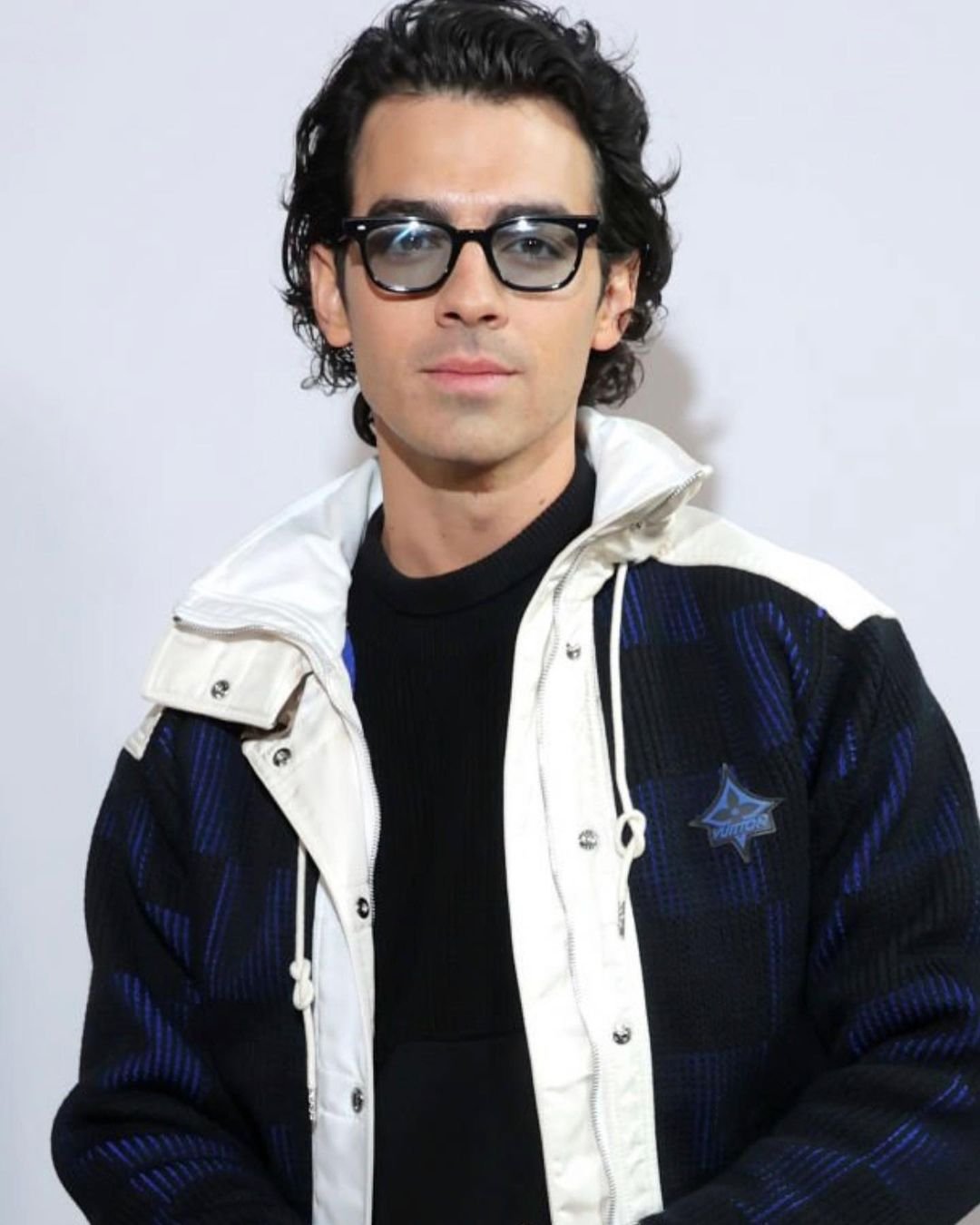 Joe Jonas wearing black glasses and a patterned jacket with white trim against a plain background