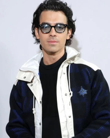 Joe Jonas wearing black glasses and a patterned jacket with white trim against a plain background