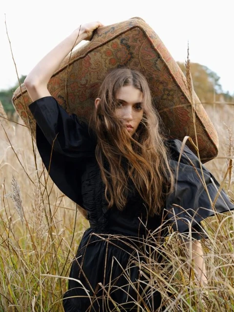 Woman in black dress carrying patterned cushion in grassy field. Portrait-style shot showcases fashion and outdoor setting.