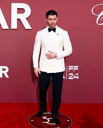 Nick Jonas in a white double-breasted tuxedo jacket and black bow tie standing on a red carpet step and repeat.