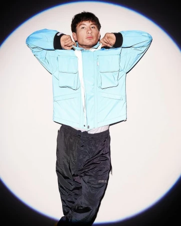Barry Keoghan wearing a light blue utility jacket and dark trousers standing in a circular spotlight against a white background.