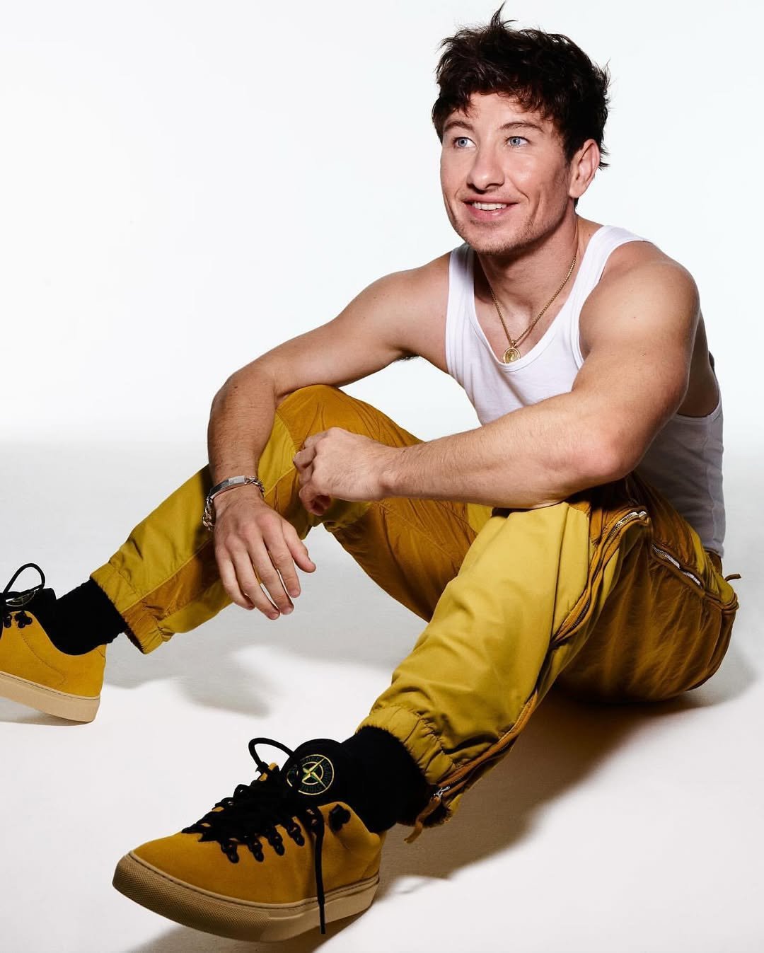 Barry Keoghan poses in a studio setting wearing a white tank top, yellow pants, and shoes with black laces and Stone Island socks.