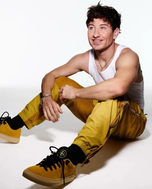 Barry Keoghan poses in a studio setting wearing a white tank top, yellow pants, and shoes with black laces and Stone Island socks.