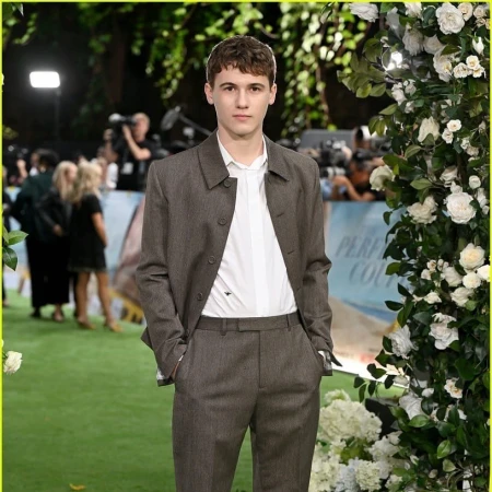 Young man standing on a green carpet wearing a brown suit jacket and matching trousers over a white shirt, with a large floral pillar beside him