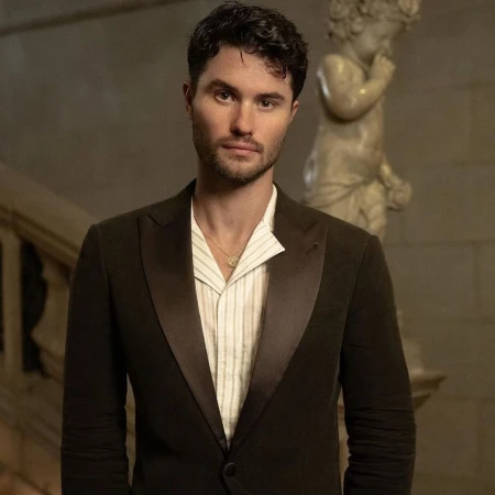 Close-up portrait of a stylish adult male in a dark tuxedo jacket and vertically striped shirt against a neutral indoor setting.