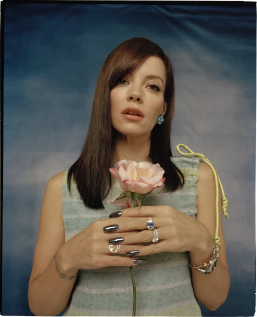 Portrait of a woman holding a pink rose in front of a sky-blue and white cloud backdrop, showcasing her unique rings and dark nail polish.