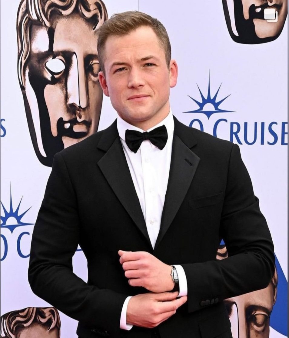 Taron Egerton, in a black tuxedo and bow tie, smiles on a red carpet at an event, with an O Cruise sign in the background.