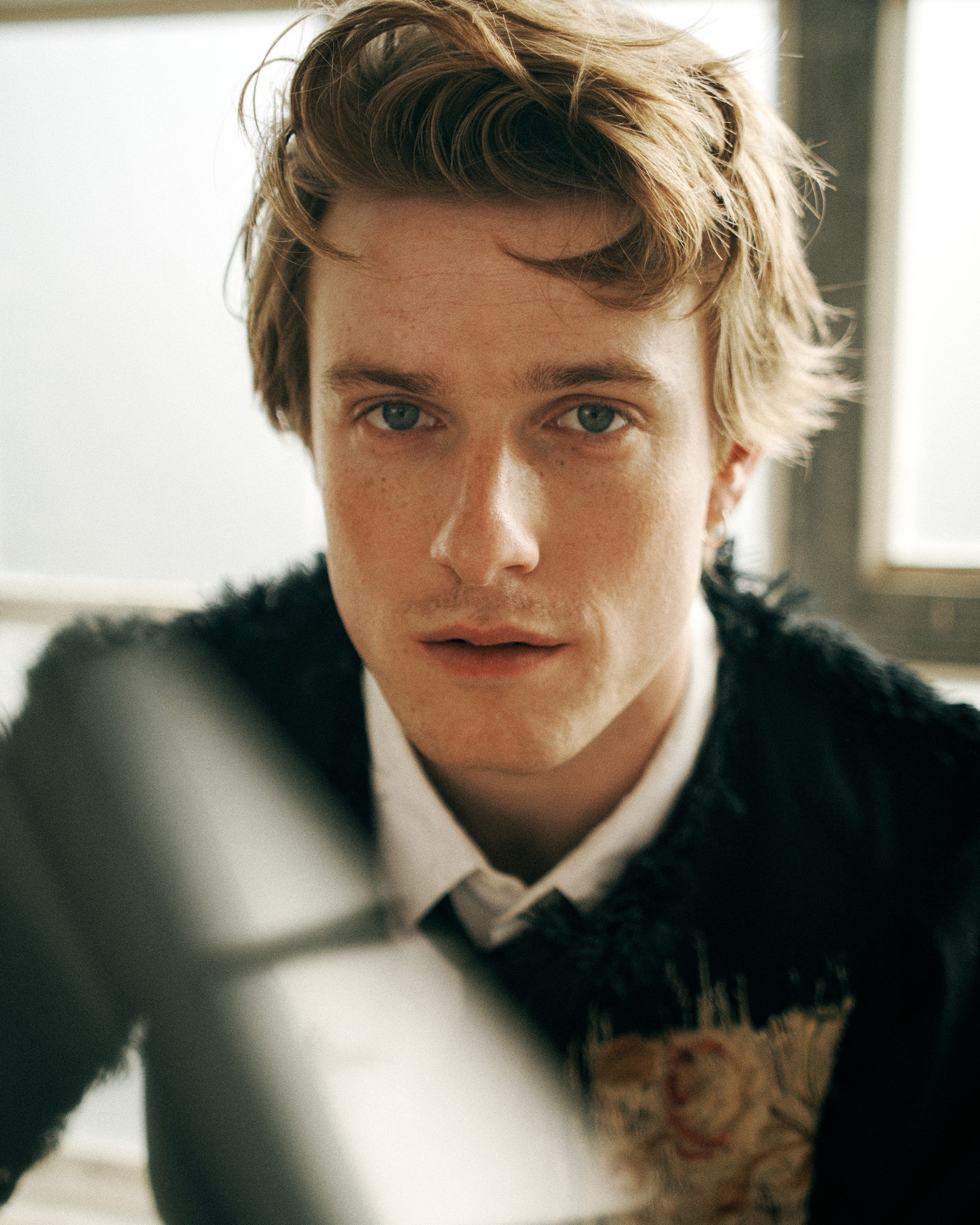 Close-up portrait of a young man with blonde hair and freckles wearing a dark textured jacket and white shirt
