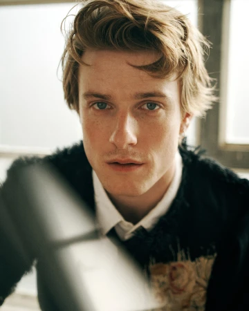 Close-up portrait of a young man with blonde hair and freckles wearing a dark textured jacket and white shirt