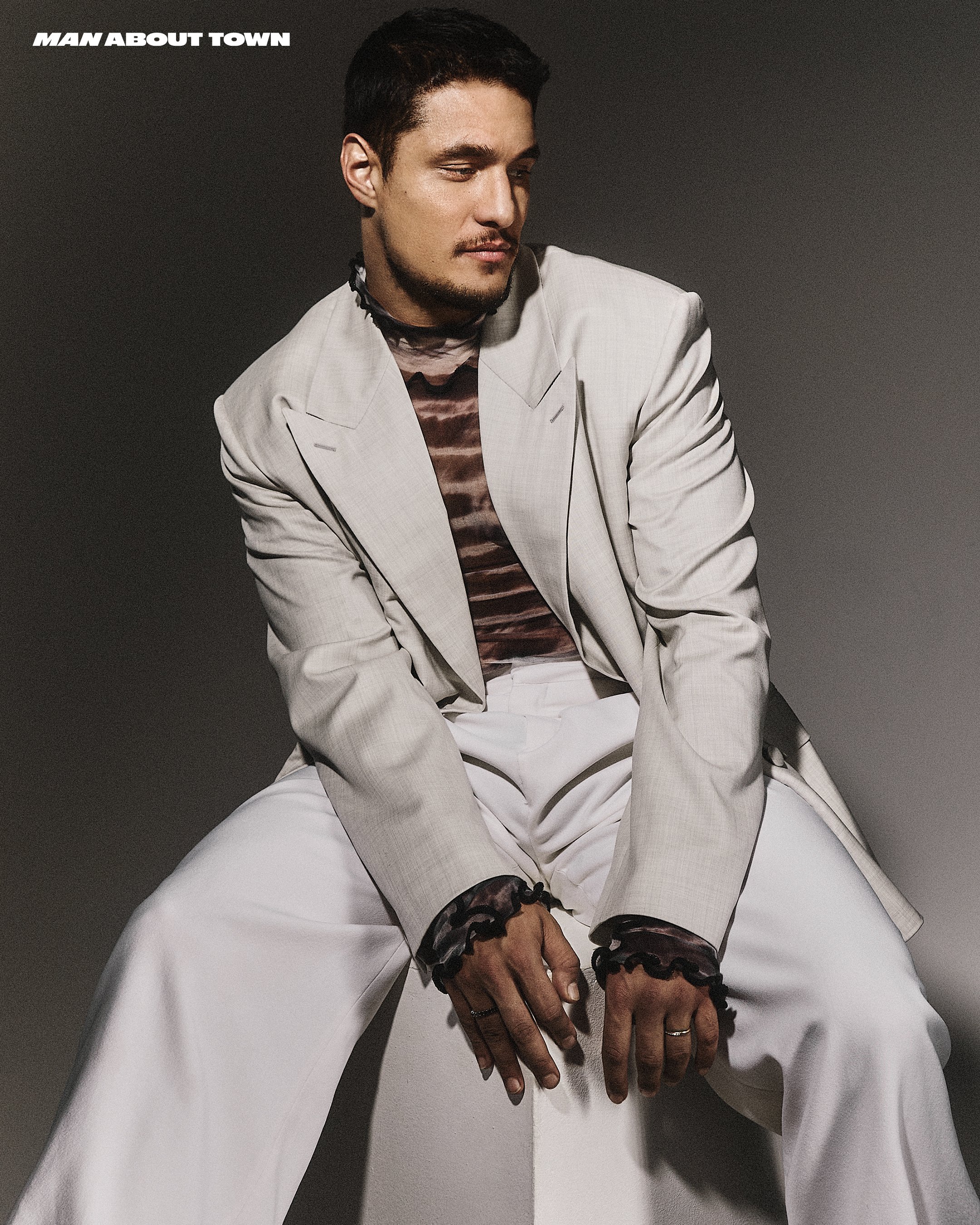Man seated wearing a light gray blazer, white trousers, and a patterned top with dark ruffled cuffs against a plain gray background.