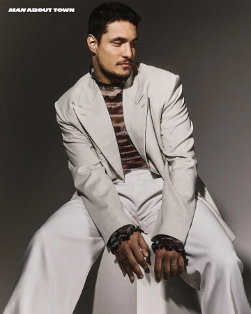 Man seated wearing a light gray blazer, white trousers, and a patterned top with dark ruffled cuffs against a plain gray background.