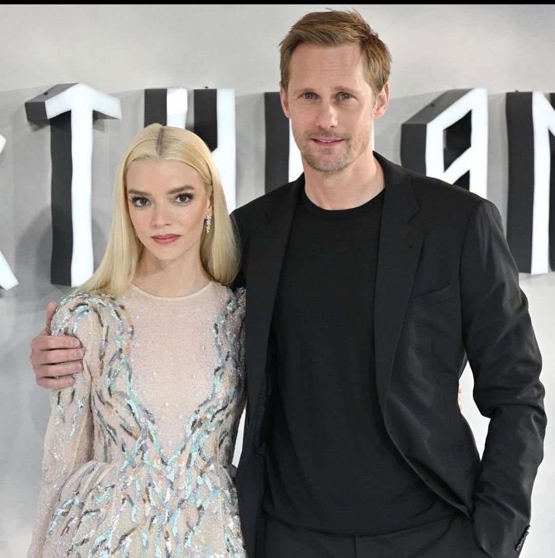 Anya Taylor-Joy and Alexander Skarsgård pose together on a red carpet, with Anya wearing a sequined dress and Alexander in a black suit.