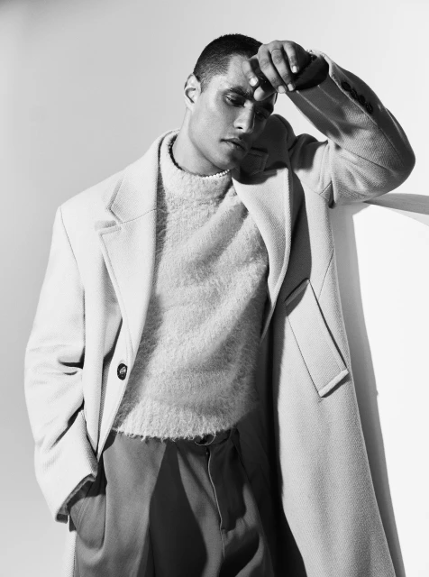 Black and white portrait of a man wearing a coat and sweater, leaning against a wall in a studio setting with soft, diffused lighting.